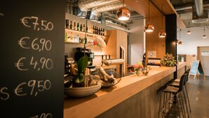 A bar area with a chalkboard menu listing prices, a variety of bottles on shelves, and a modern interior design with exposed ductwork and pendant lighting. - Hotel Trezor (Singen)