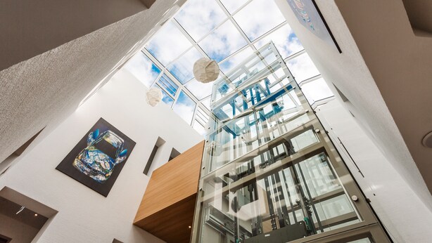 A modern interior with a glass elevator shaft, a ceiling with a geometric pattern, and a framed artwork on the wall. - Hotel Trezor (Singen)