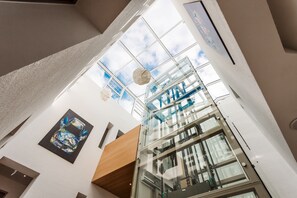 A modern interior with a glass elevator shaft, a ceiling with a geometric pattern, and a framed artwork on the wall. - Hotel Trezor (Singen)