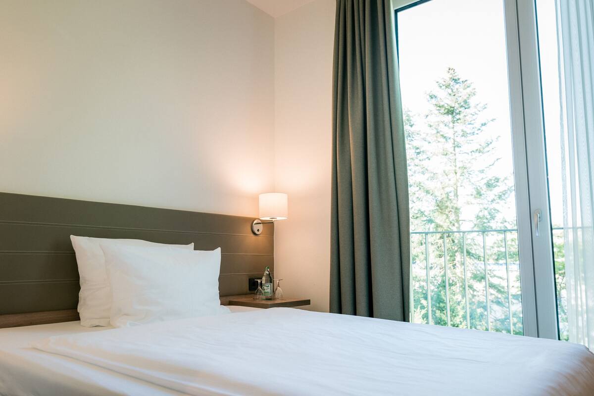 A hotel room with a neatly made bed, a bedside lamp, a nightstand, and a window with curtains.