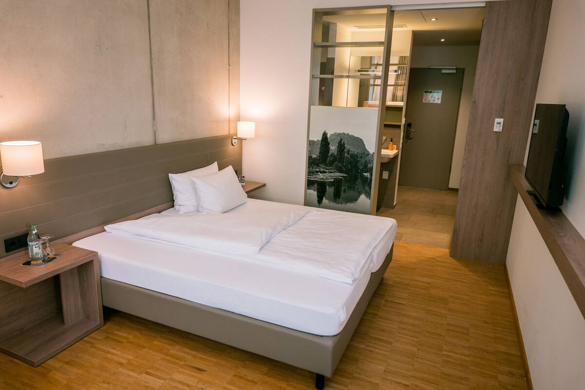 A hotel room with a bed, bedside lamps, a nightstand, a TV, and a wooden floor.