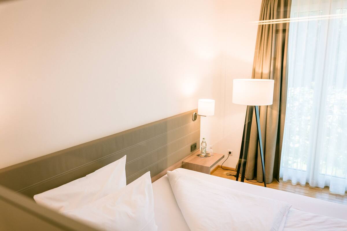 A hotel room with a bed, a nightstand, a lamp, and a window with curtains.