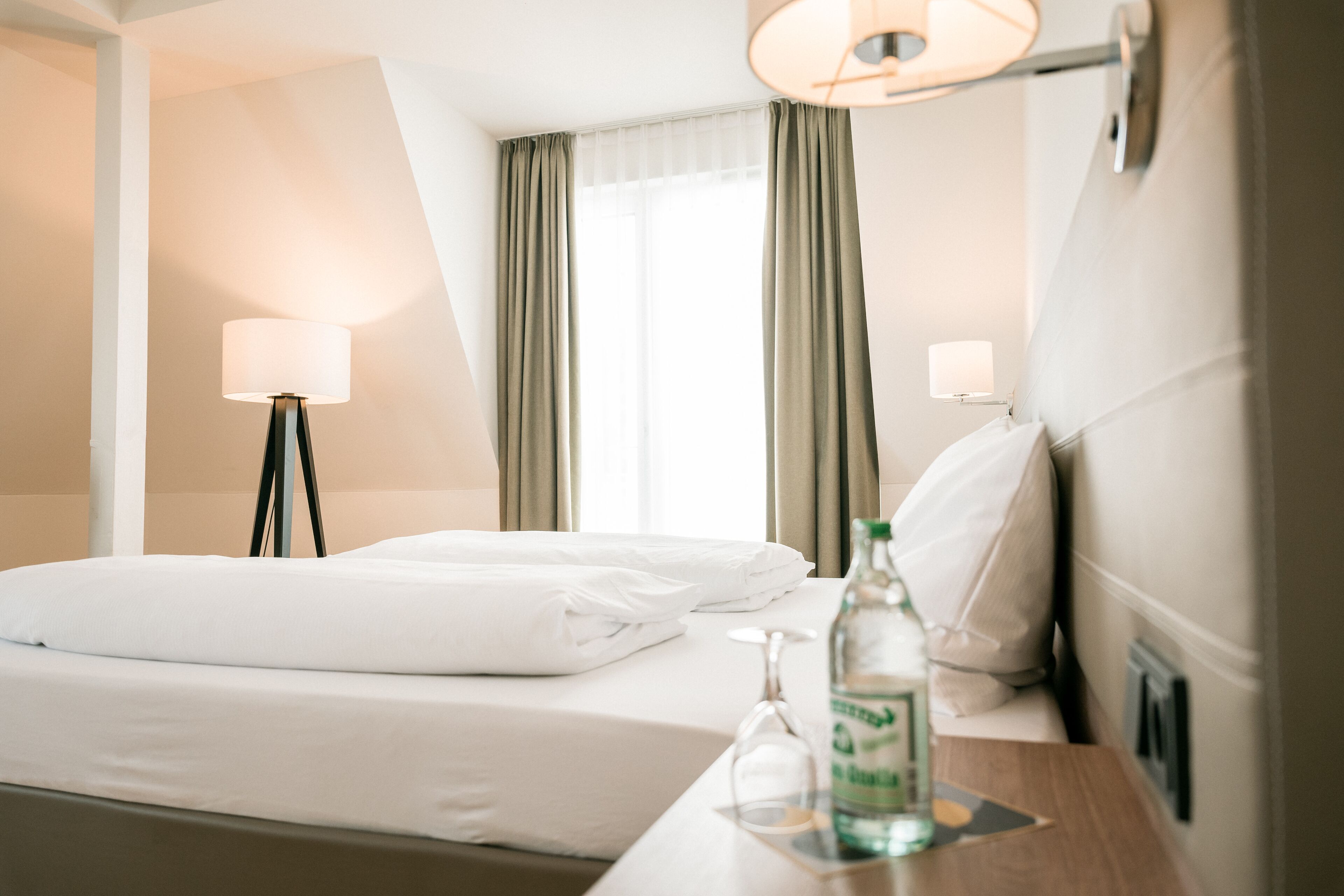 A hotel room with a neatly made bed, a bedside table with a bottle and glass, and a lamp.