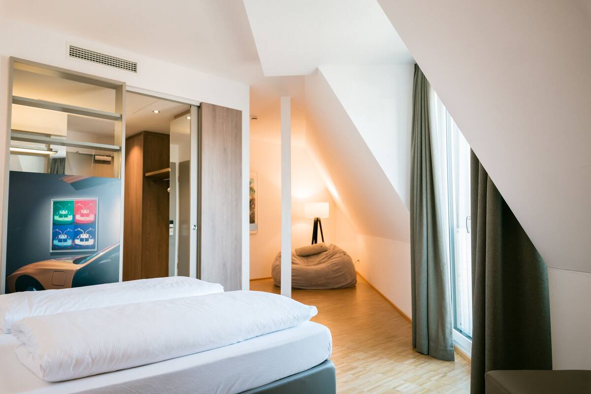 A modern hotel room with a bed, a desk, a bean bag, and a large window with curtains.