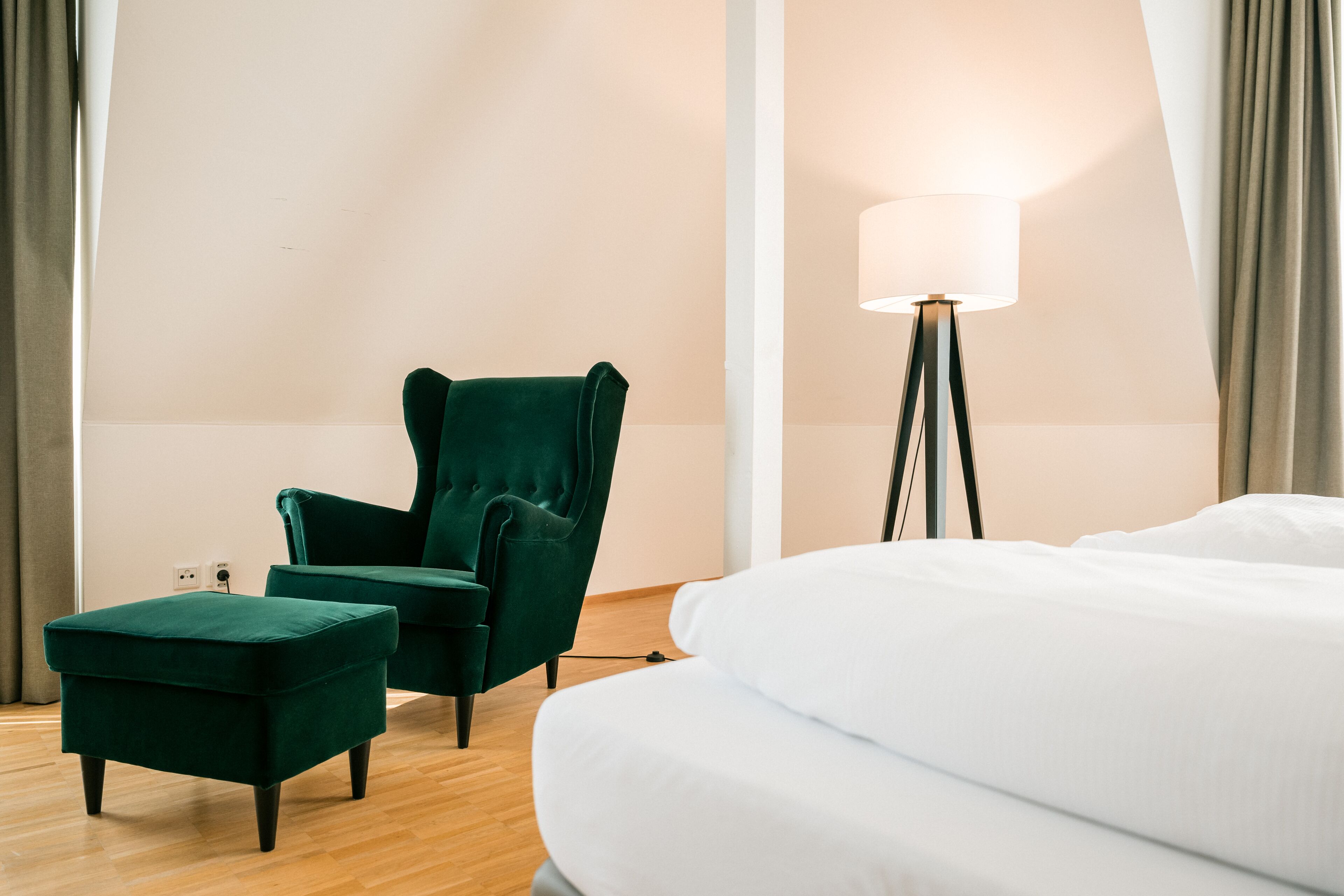 Une chambre d’hôtel avec un lit, un fauteuil vert, une lampe sur pied et un petit pouf.