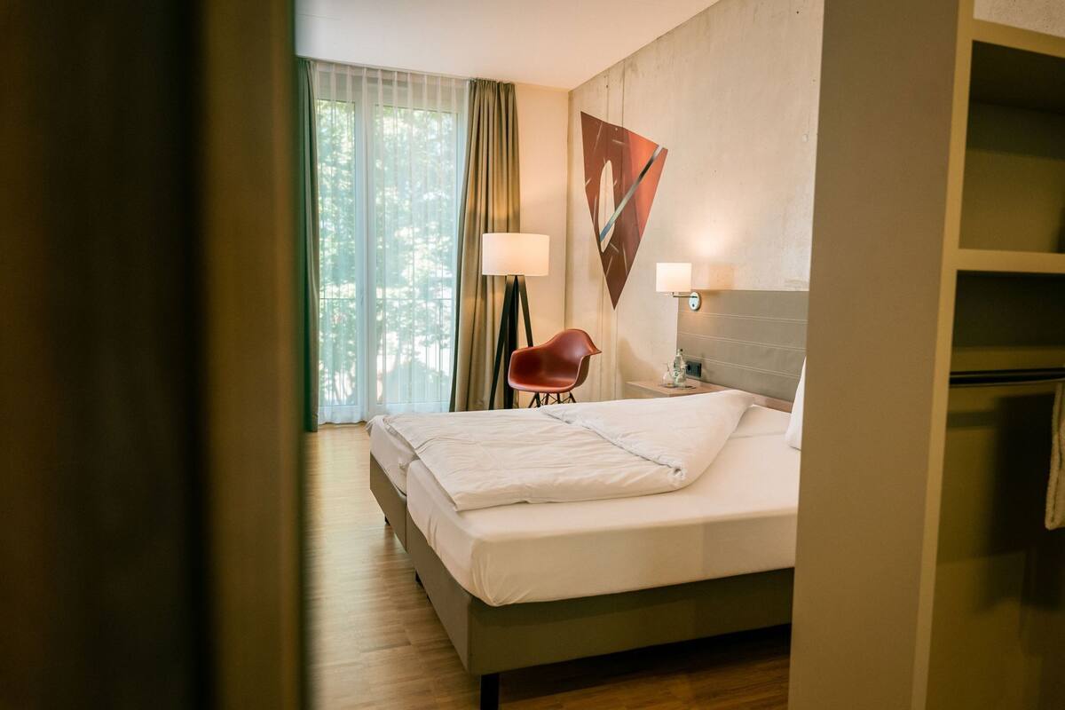 A hotel room with a bed, a chair, a lamp, and a painting on the wall.