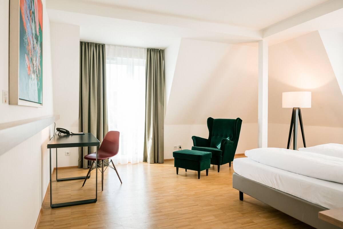 A modern hotel room with a bed, desk, two chairs, and a lamp.