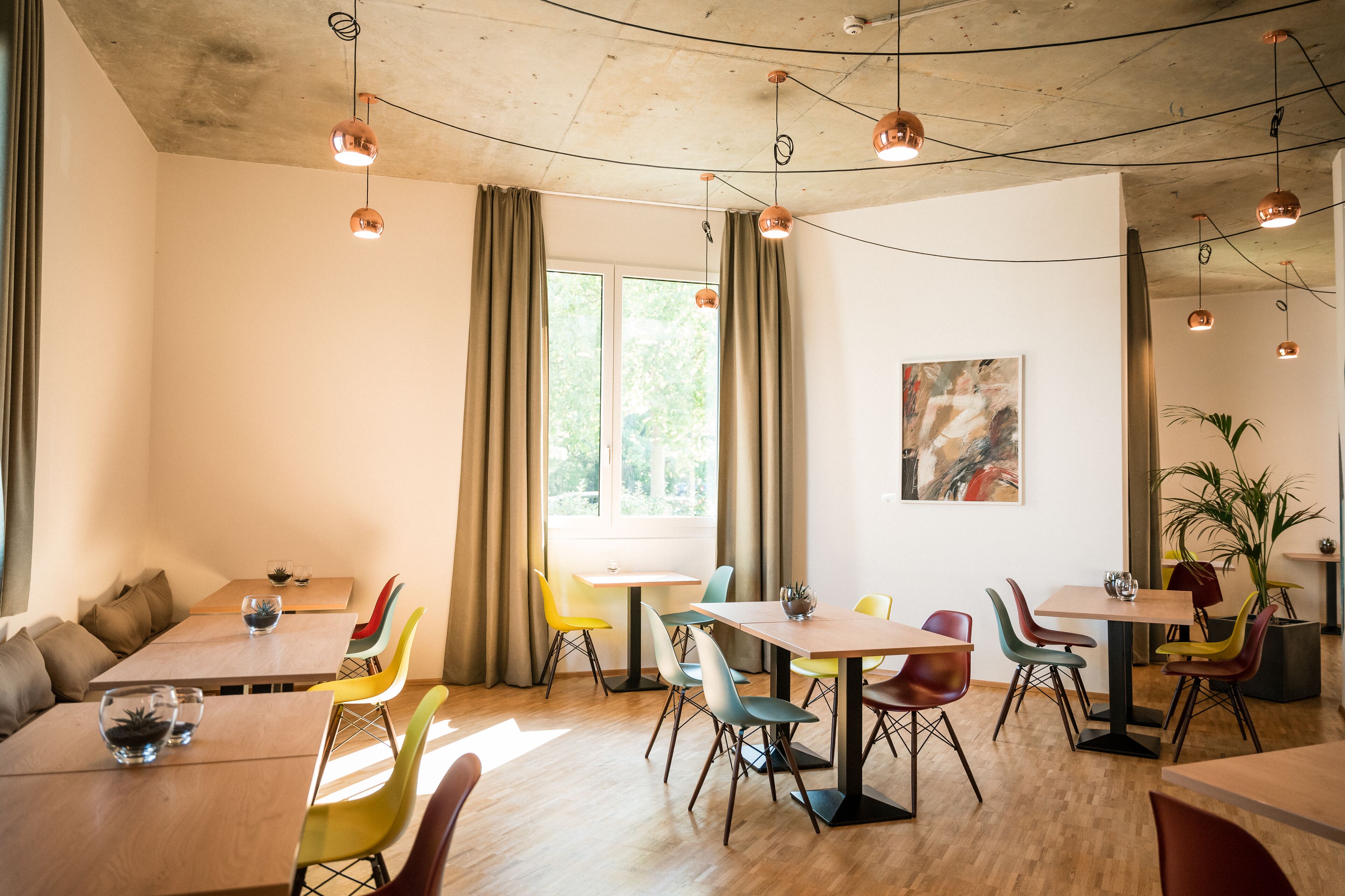 A modern dining area with wooden tables and colorful chairs, large windows with curtains, and pendant lighting.