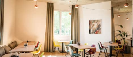 A modern dining area with wooden tables and colorful chairs, large windows with curtains, and pendant lighting.