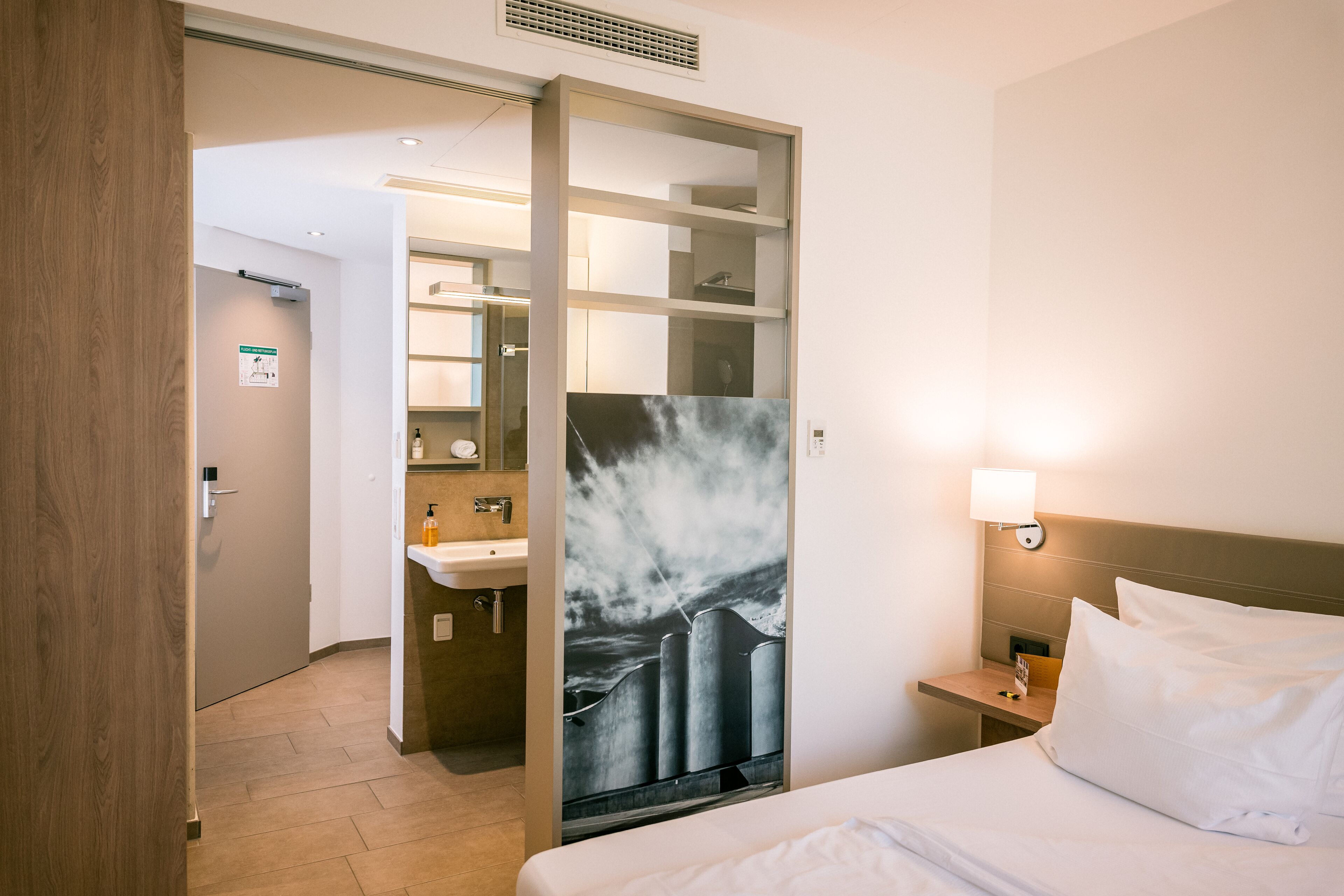 A hotel room with a bed, a nightstand, a wall-mounted lamp, and a bathroom visible through an open door.
