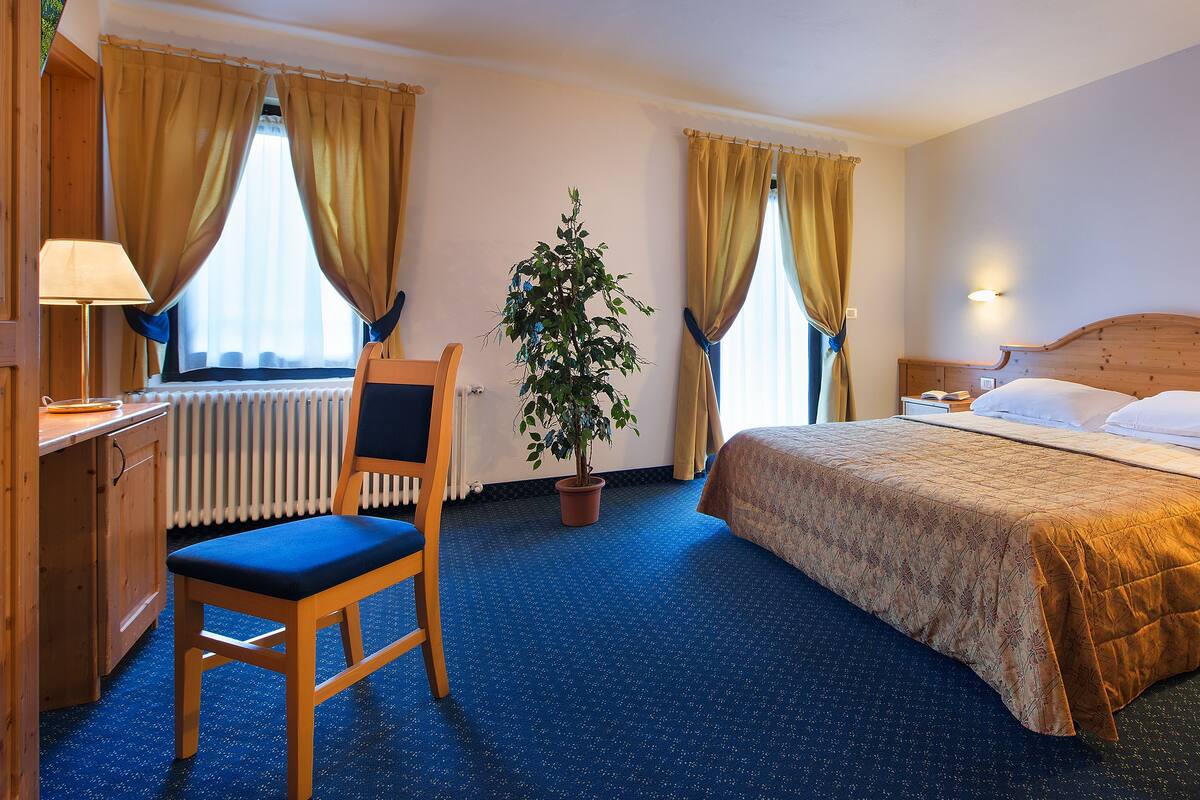 Habitación de hotel con cama, mesita de noche, lámpara, sillón, planta en maceta y escritorio con lámpara.
