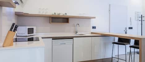 Une cuisine moderne avec des meubles blancs, un four à micro-ondes encastré, un lave-vaisselle et un plan de travail en bois. On y trouve deux tabourets de bar noirs et une étagère en bois au-dessus de l’évier.