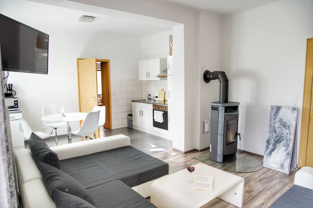 A modern living room with a sofa, a dining area with a table and chairs, a kitchen with cabinets and a stove, and a fireplace.
