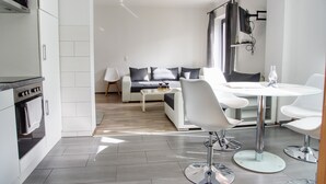 A modern kitchen with white cabinets and a built-in oven, a dining area with a glass table and white chairs, and a living area with a sofa and coffee table. - Haff-Ostseeferienhaus (Monkebude)