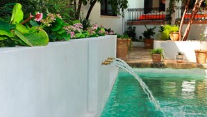 Piscina com água jorrando de uma torneira fixada na parede, rodeada por plantas em vasos e um muro branco.