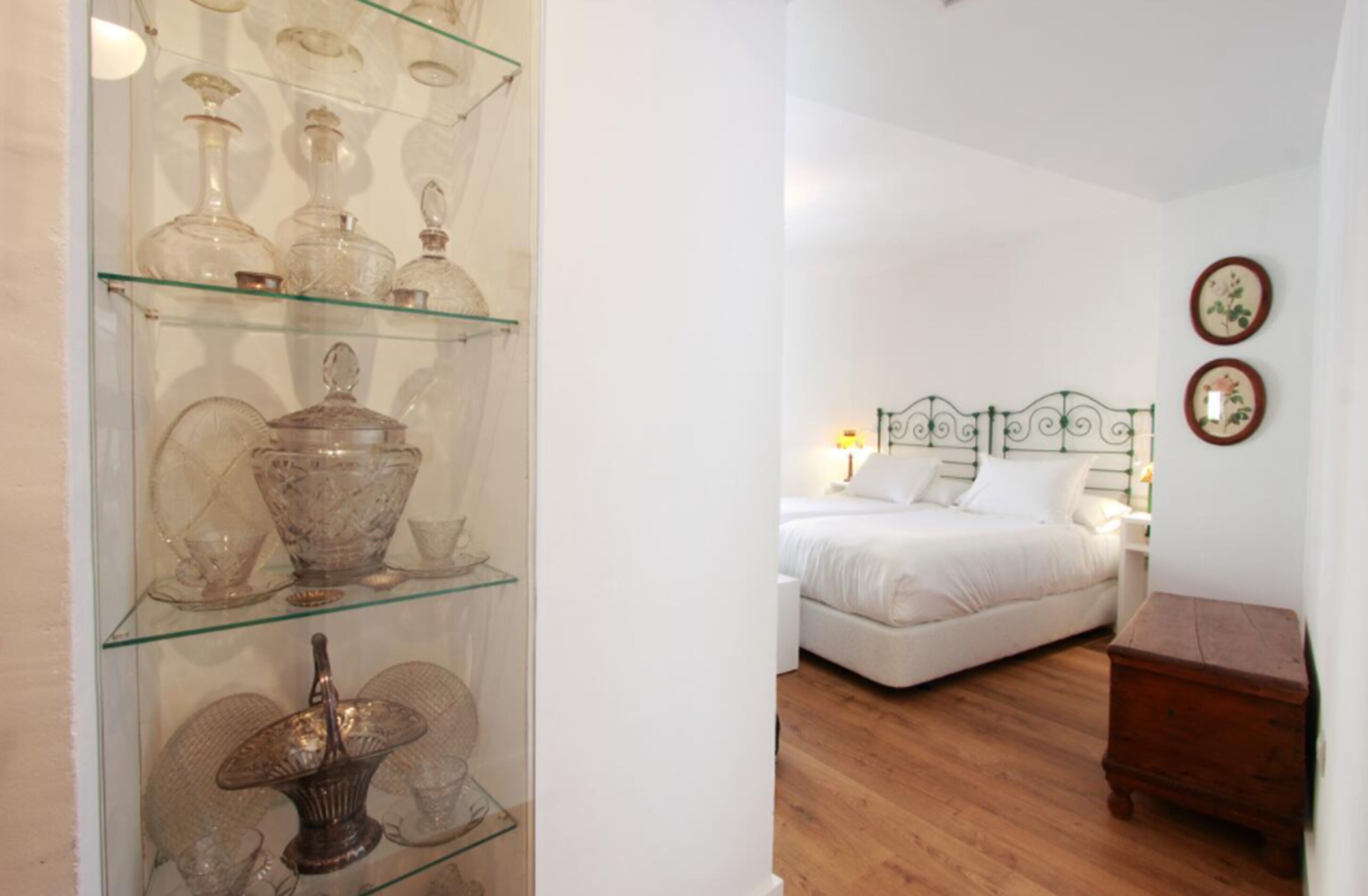 Un dormitorio con cama, mesita de noche y tocador. A la izquierda, un expositor de vidrio con diversos vasos.