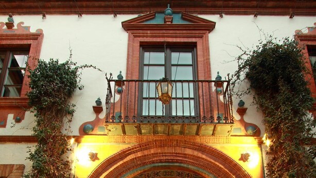 Uma entrada imponente com um arco trabalhado, uma grande fonte decorativa e vegetação exuberante.