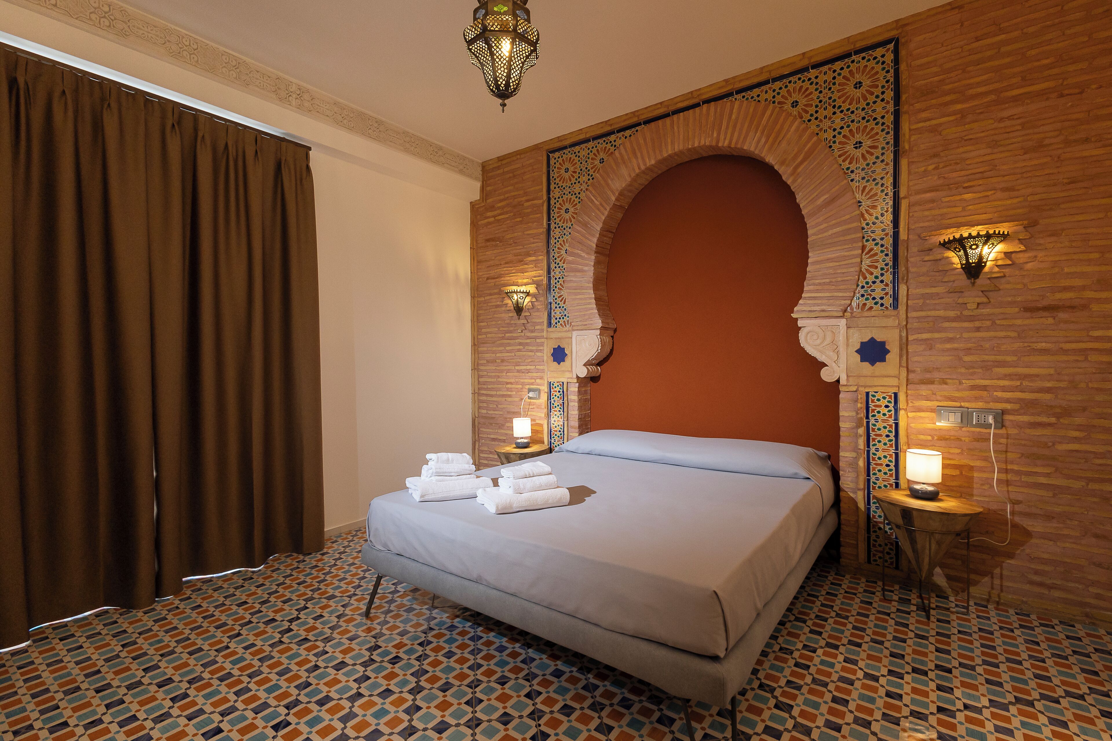 A hotel room with a bed, a nightstand, a wall-mounted lamp, and a patterned floor.