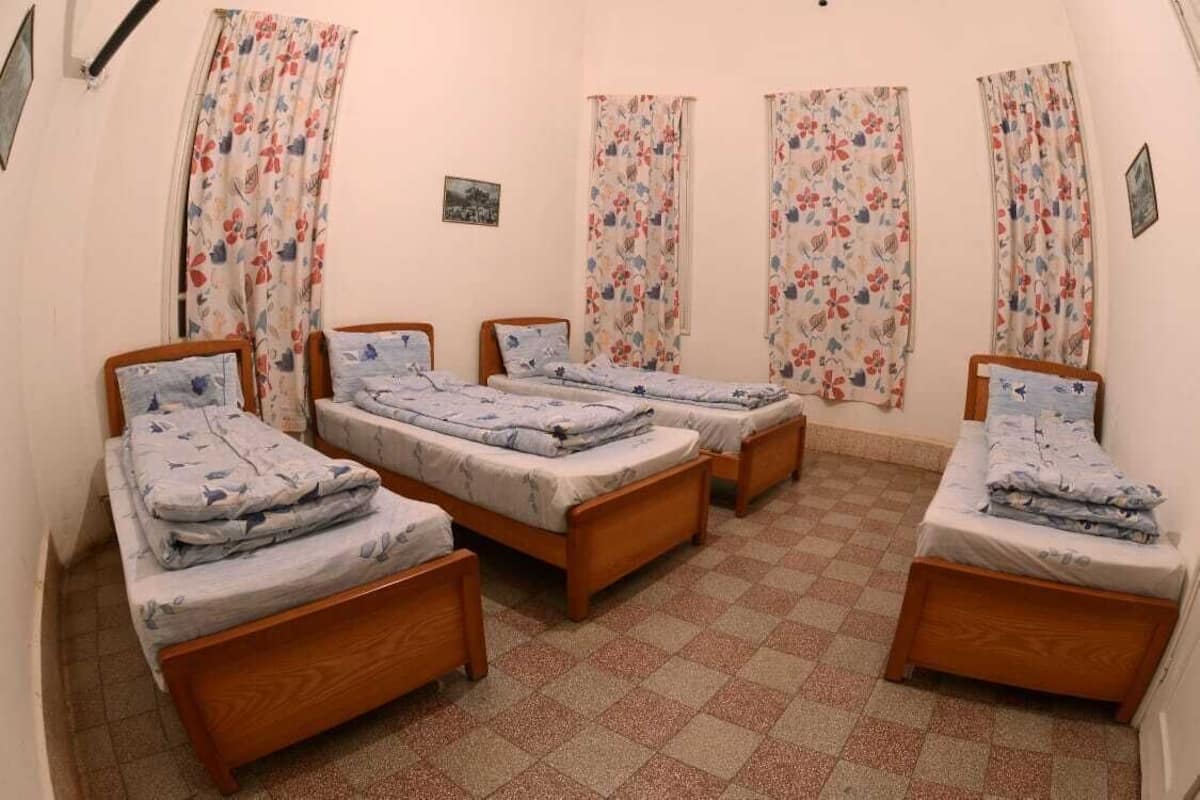Une chambre avec quatre lits simples, chacun doté d’un cadre en bois et d’une couvre-lit à motifs floraux. La chambre a un sol à carreaux et est agrémentée de rideaux à motifs floraux aux fenêtres.