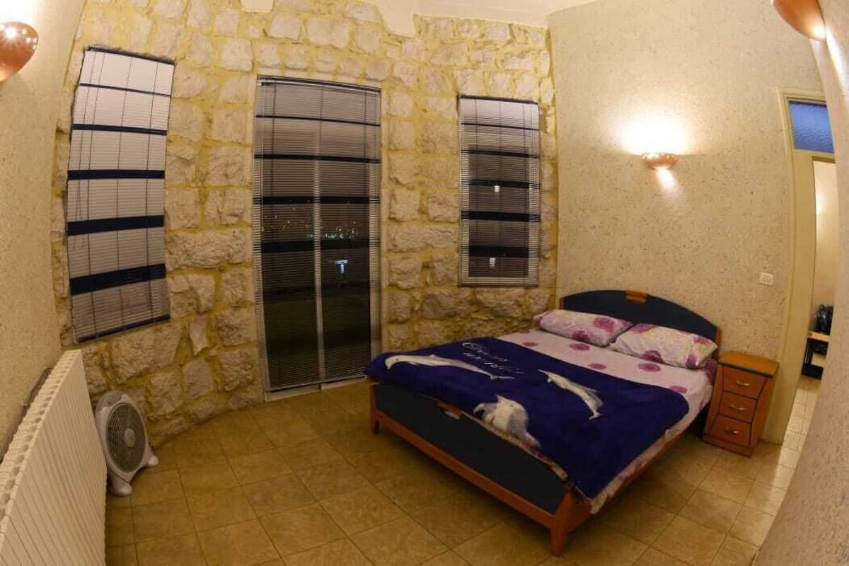 Une chambre avec un mur en pierre, un lit recouvert d’une courtepointe bleue et blanche, une table de chevet et une fenêtre avec des stores.