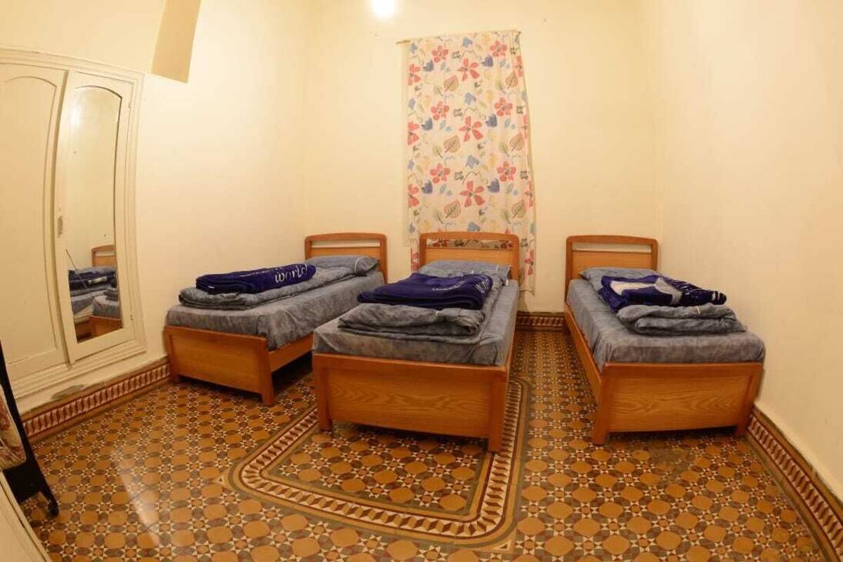 Une pièce avec trois lits en bois disposés en rangée, un miroir, un sol à motifs et un rideau à fleurs.