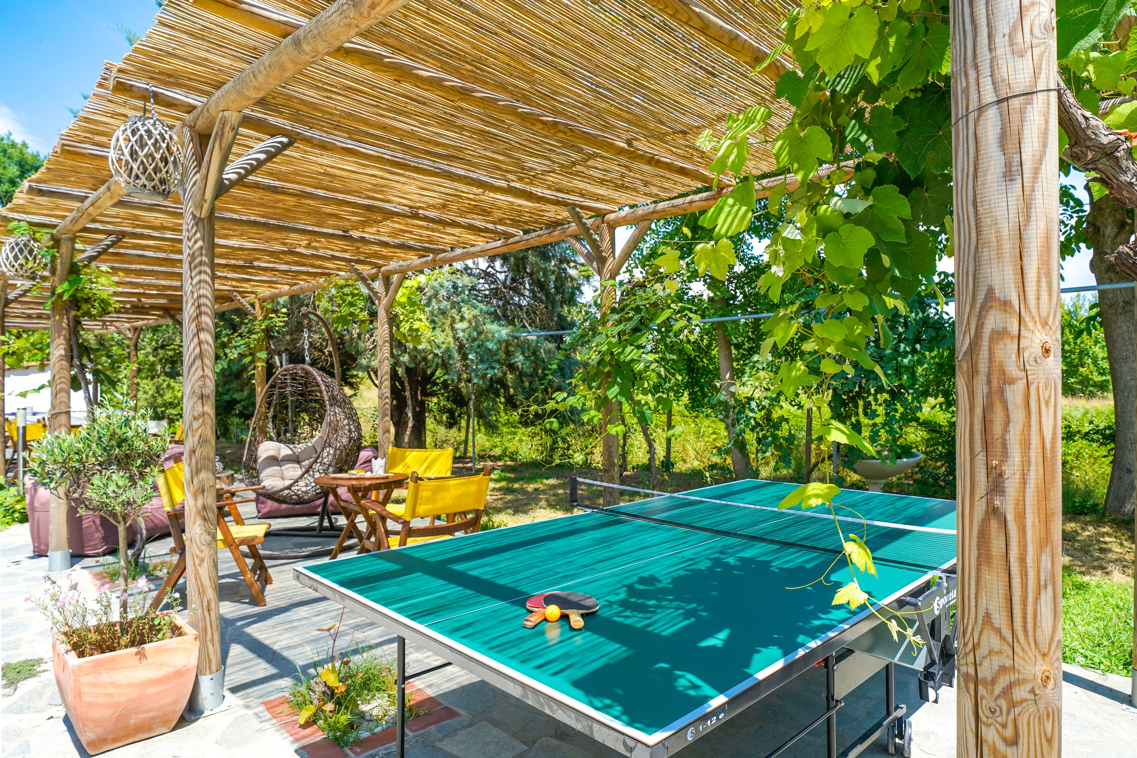 An outdoor ping pong table under a wooden pergola with a hanging ball, surrounded by potted plants and outdoor furniture.