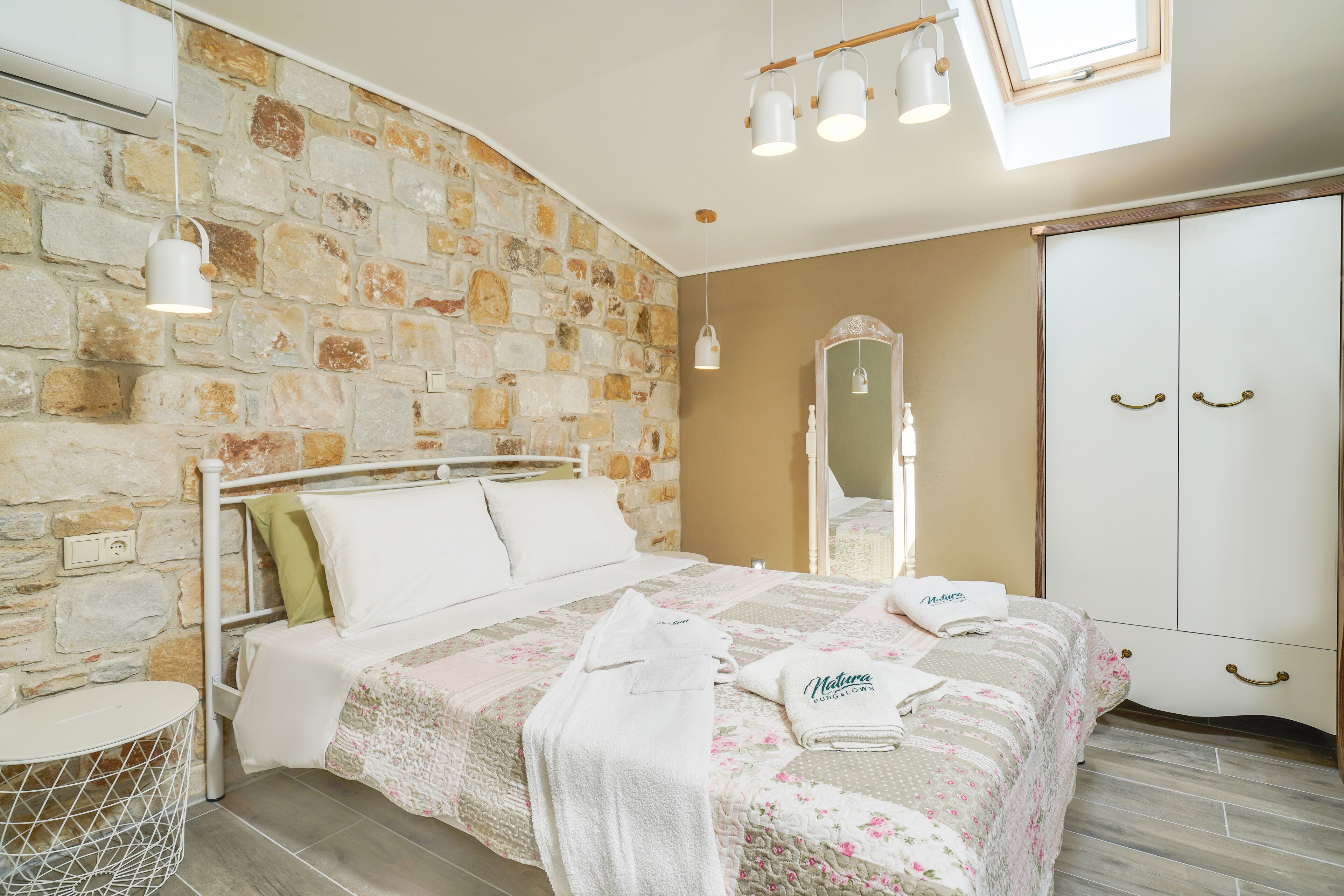 A bedroom with a stone wall, a bed with floral bedding, a round bedside table, and a wardrobe with a mirror.