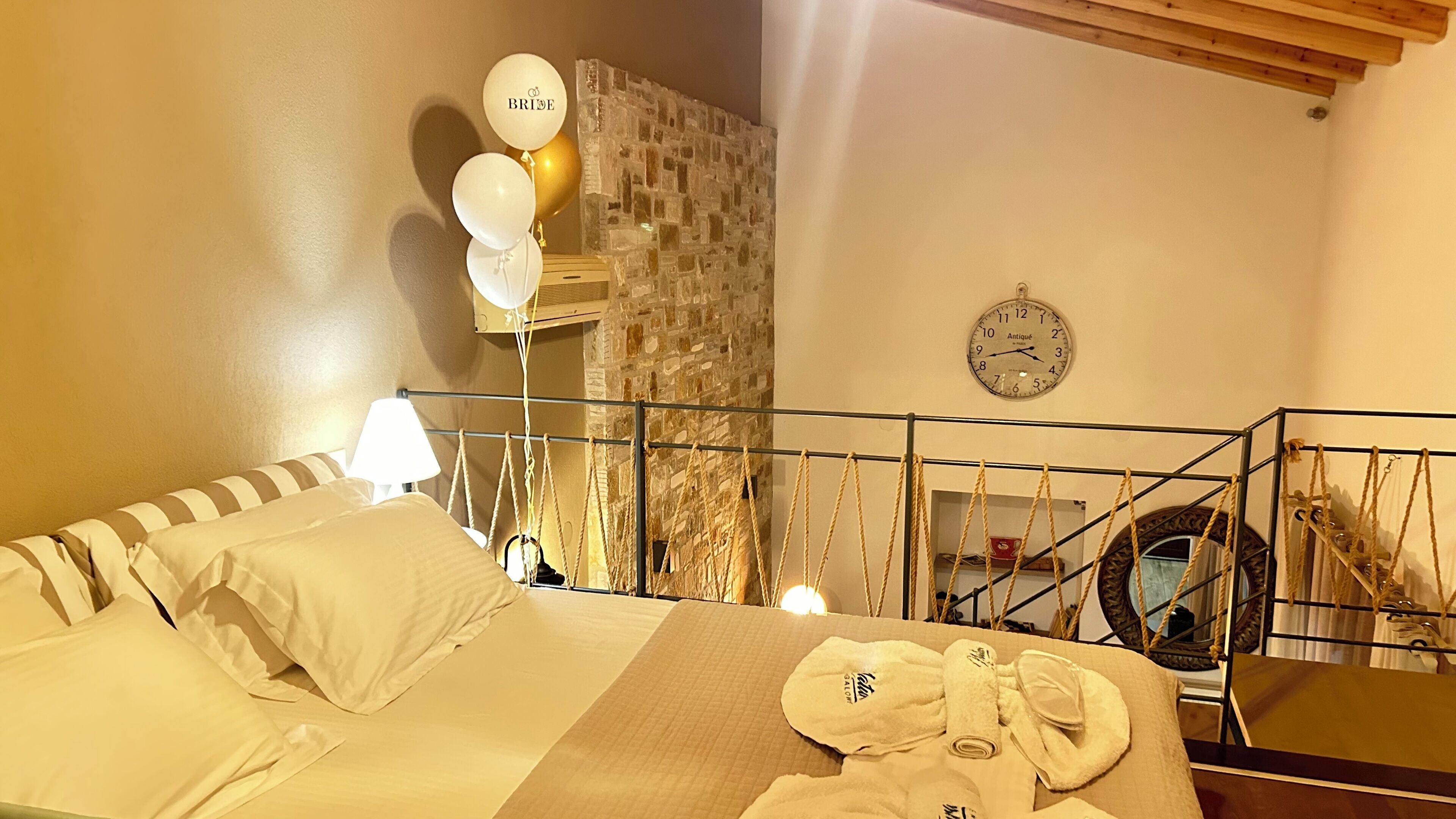 A hotel room with a bed, a stone wall, a clock, and a balcony with ropes.