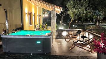 A hot tub with a stone wall and wooden deck, surrounded by outdoor seating and a pool.