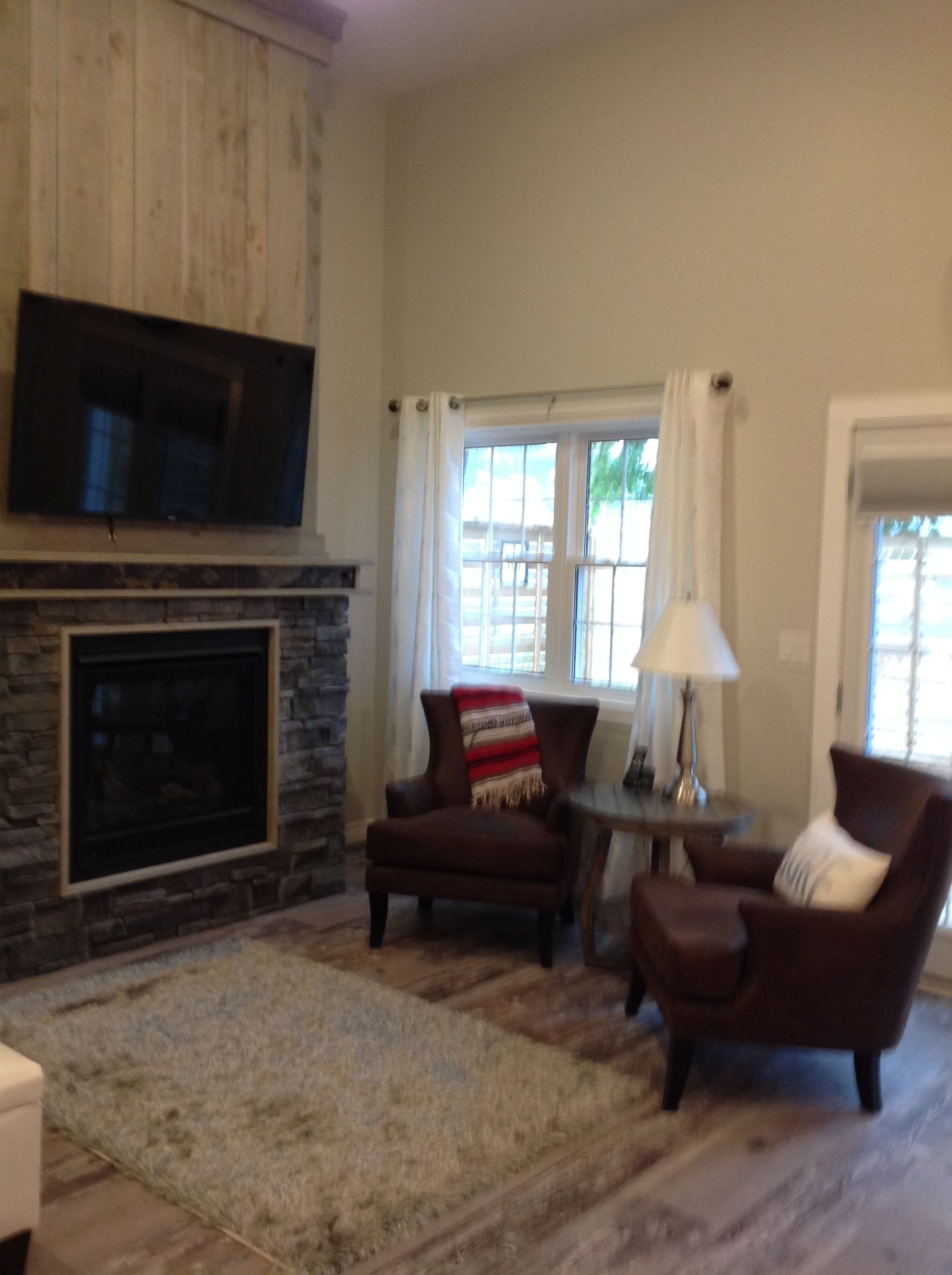 A living room with a stone fireplace, two brown armchairs, a small round table with a lamp, and a television mounted on the wall.