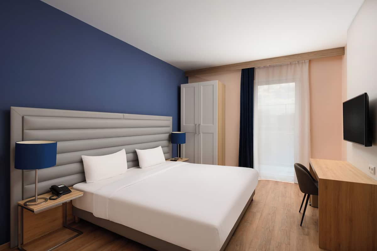Une chambre d’hôtel avec un grand lit, des tables de chevet, un bureau, une chaise, une télévision et une fenêtre avec des rideaux.