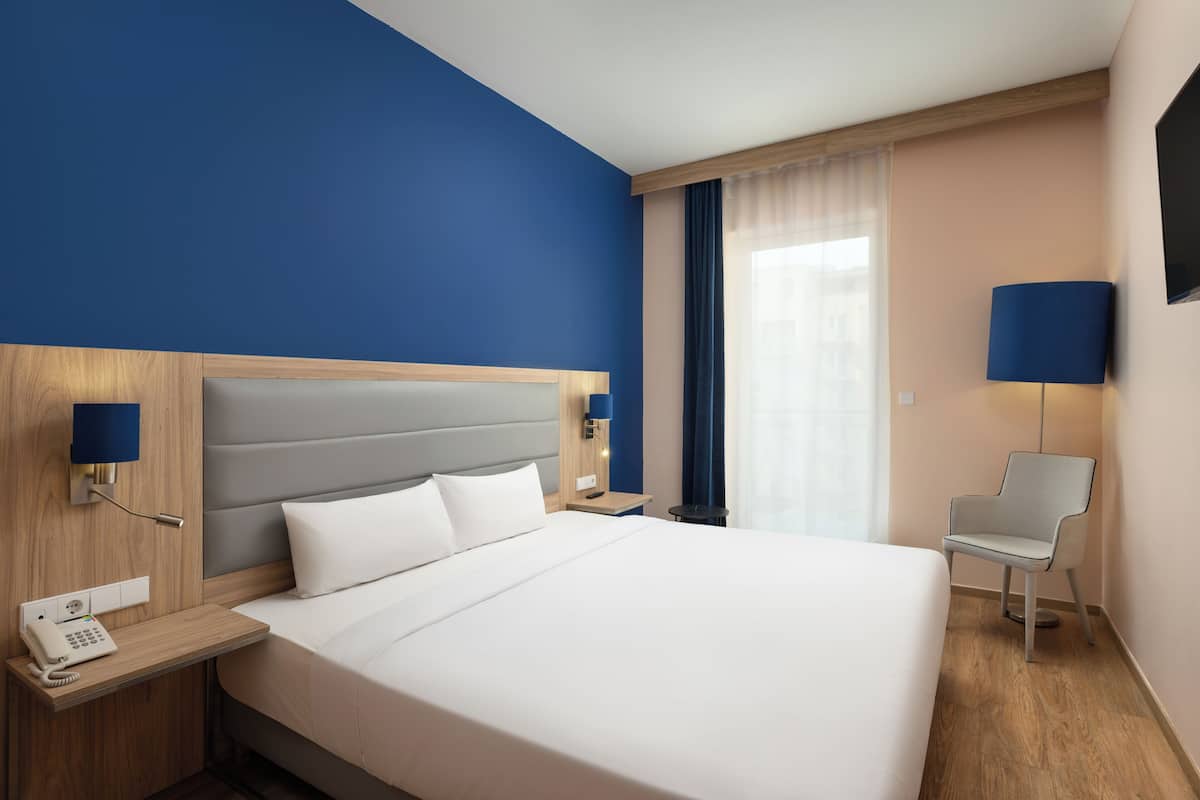 Une chambre d’hôtel avec un grand lit, un téléphone sur la table de chevet, une table de chevet, une chaise et un téléviseur fixé au mur.
