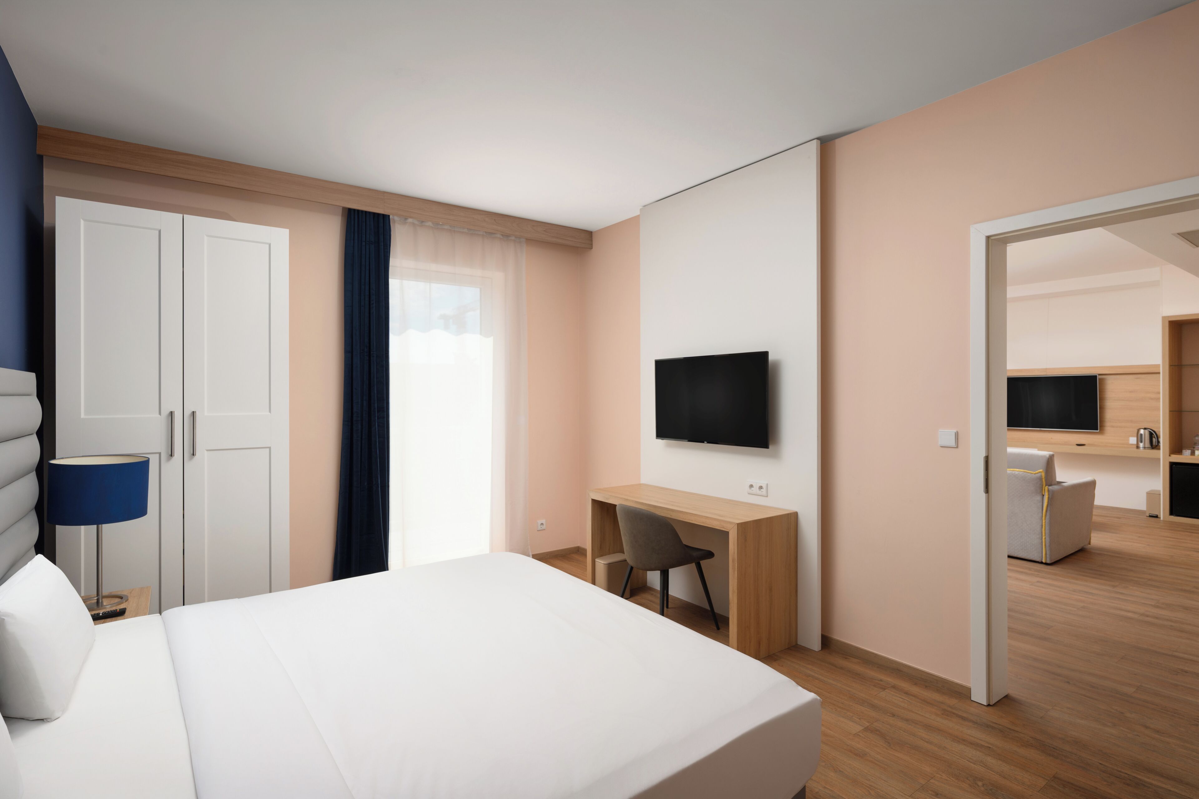 A hotel room with a bed, desk, chair, TV, wardrobe, and a view of the adjacent kitchen area.