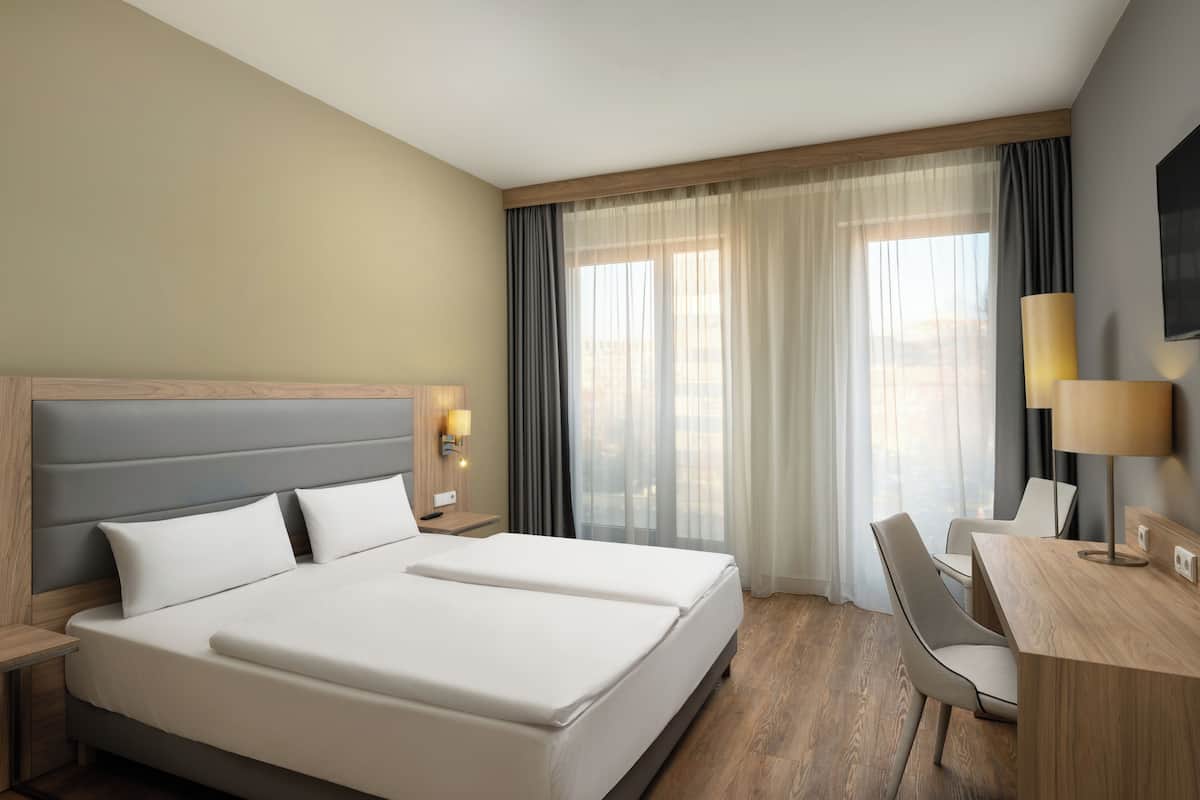 Une chambre d’hôtel avec un grand lit, un bureau avec une chaise, une télévision et une fenêtre avec des rideaux.