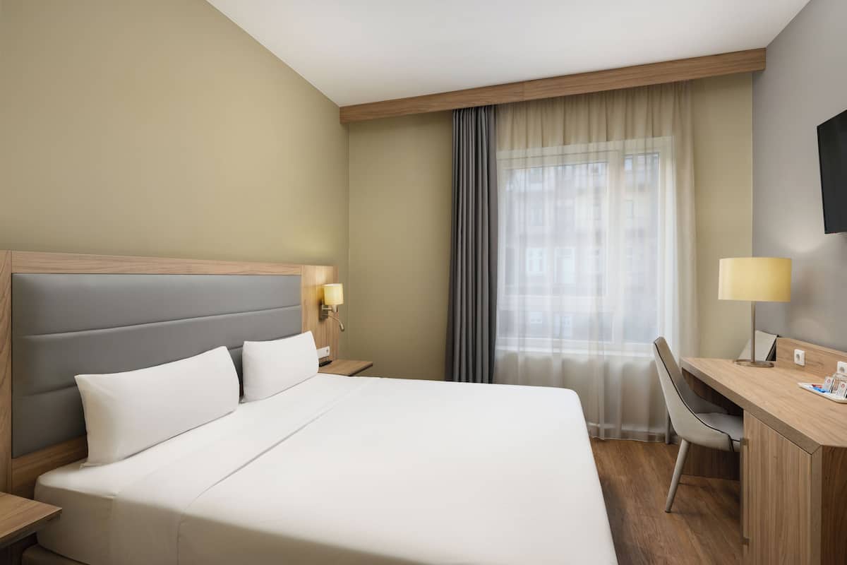 Une chambre d’hôtel avec un grand lit, un bureau avec une chaise, un téléviseur fixé au mur et une fenêtre avec des rideaux.