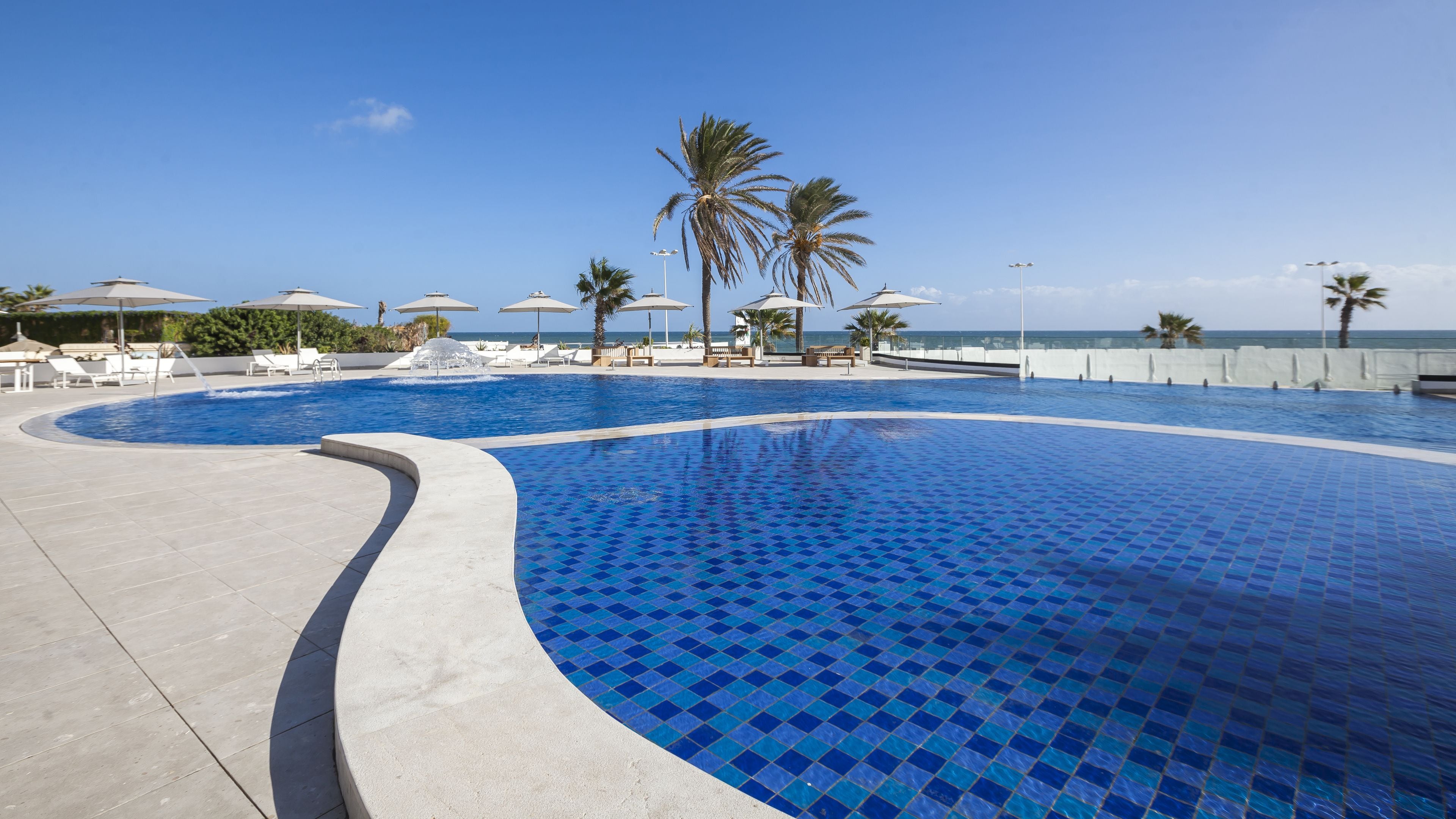 A swimming pool with a tiled edge, surrounded by lounge chairs and umbrellas, with palm trees and a clear sky in the background.