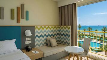 Habitación de hotel con cama, sofá, una mesita, teléfono y vistas a una piscina y playa.