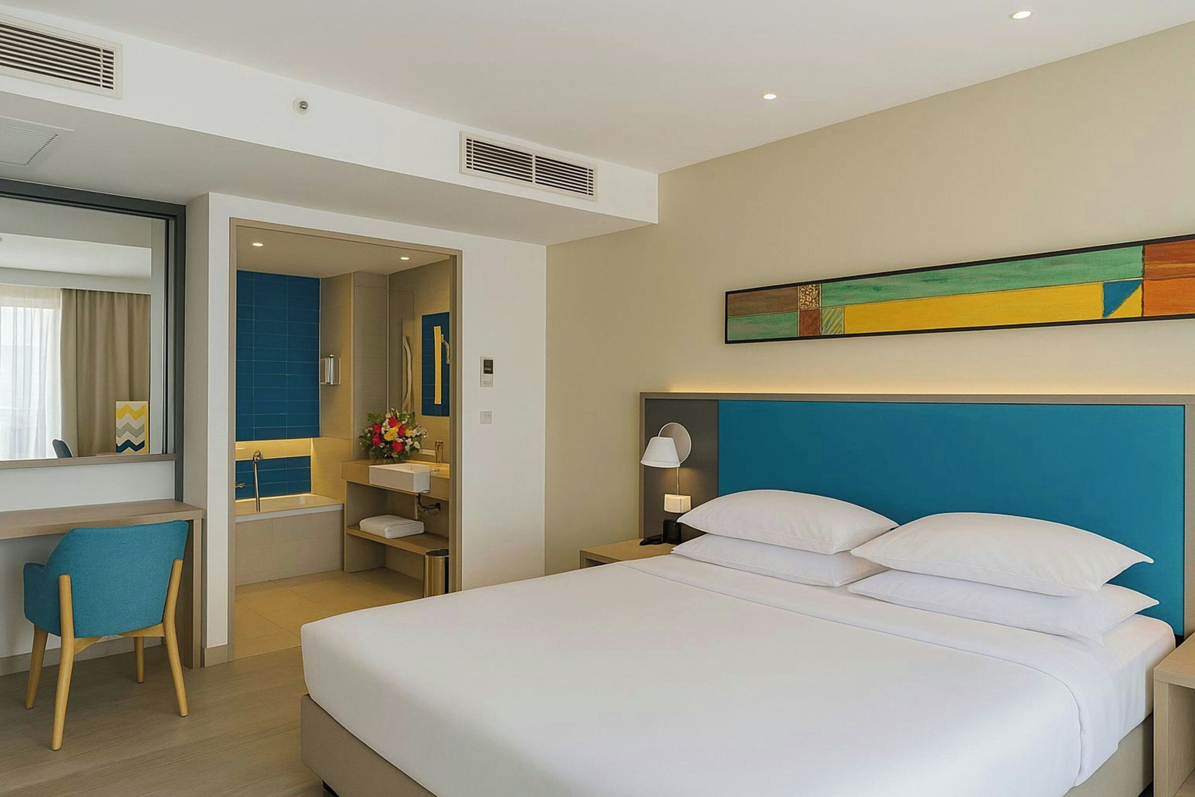 Une chambre d’hôtel avec un lit, un bureau, une chaise, une salle de bain et un tableau au mur.