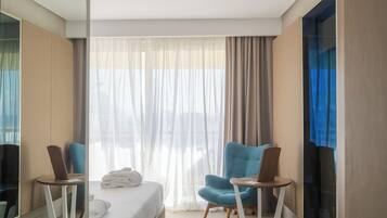 A modern hotel room with a bed, two armchairs, a small table, a mirror, and a large window with curtains.