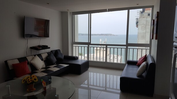 A modern living room with a large window overlooking a body of water, a flat-screen TV mounted on the wall, and a black leather sofa with red and white cushions. - Apartamento Superior Laguito (Cartagena)