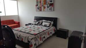 Quarto com cama, sofá vermelho, ventoinha preta, mesa de cabeceira e uma obra de arte na parede.