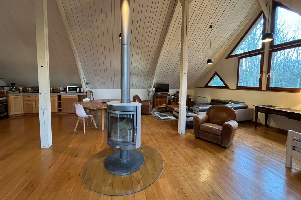 A spacious loft with a central wood-burning stove, wooden floors, and large windows offering views of the outdoors.