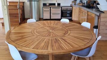Une table ronde en bois, entourée de chaises, un coin cuisine avec des placards et des appareils électroménagers, et un escalier.