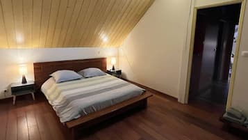 A bedroom with a wooden ceiling, a bed with striped bedding, two bedside tables with lamps, and a door leading to another room.