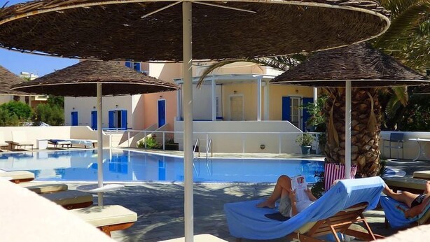 A pool area with lounge chairs and umbrellas, a clear blue pool, and a building with blue doors in the background. - Avra Hotel (Santorini)