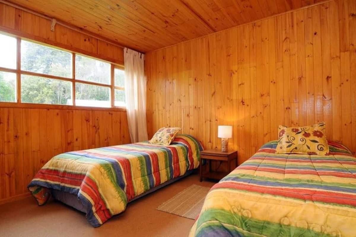 A room with wooden walls, two beds with colorful striped bedding, a window with curtains, and a small bedside table with a lamp.