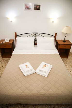 A neatly made bed with a quilted beige bedspread, two folded white towels with orange accents, a bedside lamp, and a nightstand with a telephone. - Hotel Anixis (Lamia)