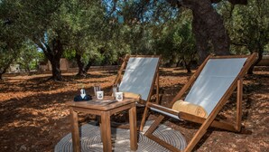 Dos sillones de madera con cojines blancos, una pequeña mesa de madera y un tapete tejido sobre un terreno de tierra rodeado de olivos.