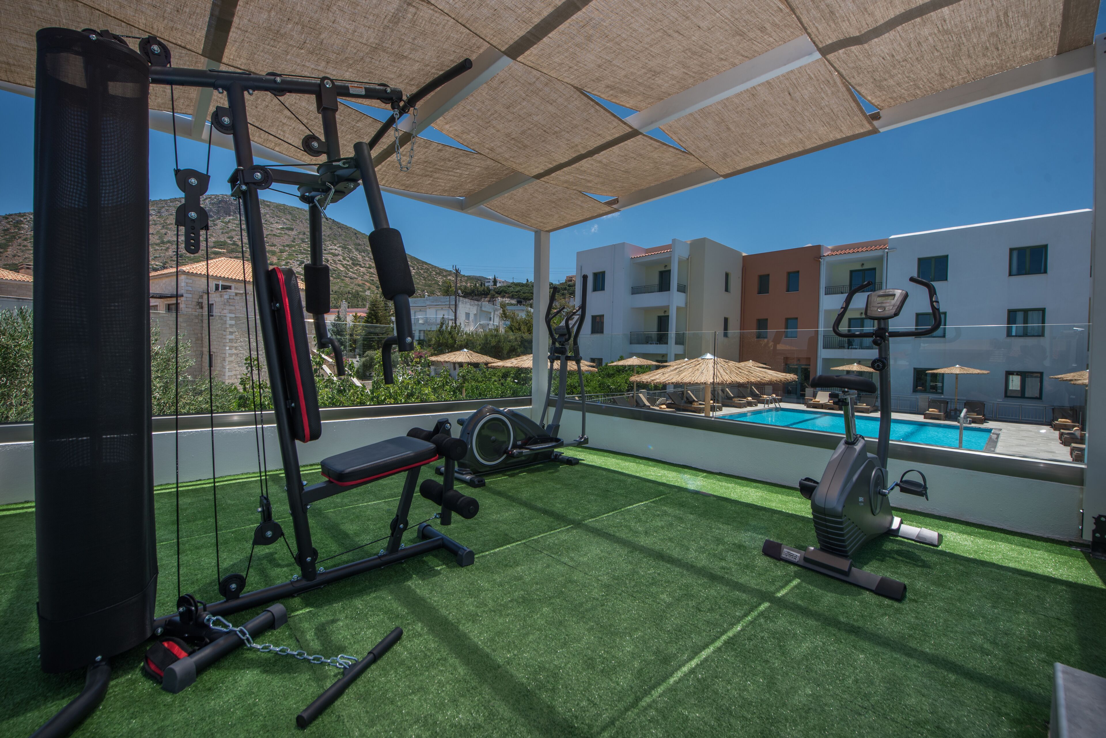 Une salle de sport située sur le toit, équipée de matériel d’exercice, avec une piscine et une vue sur les bâtiments et les montagnes.