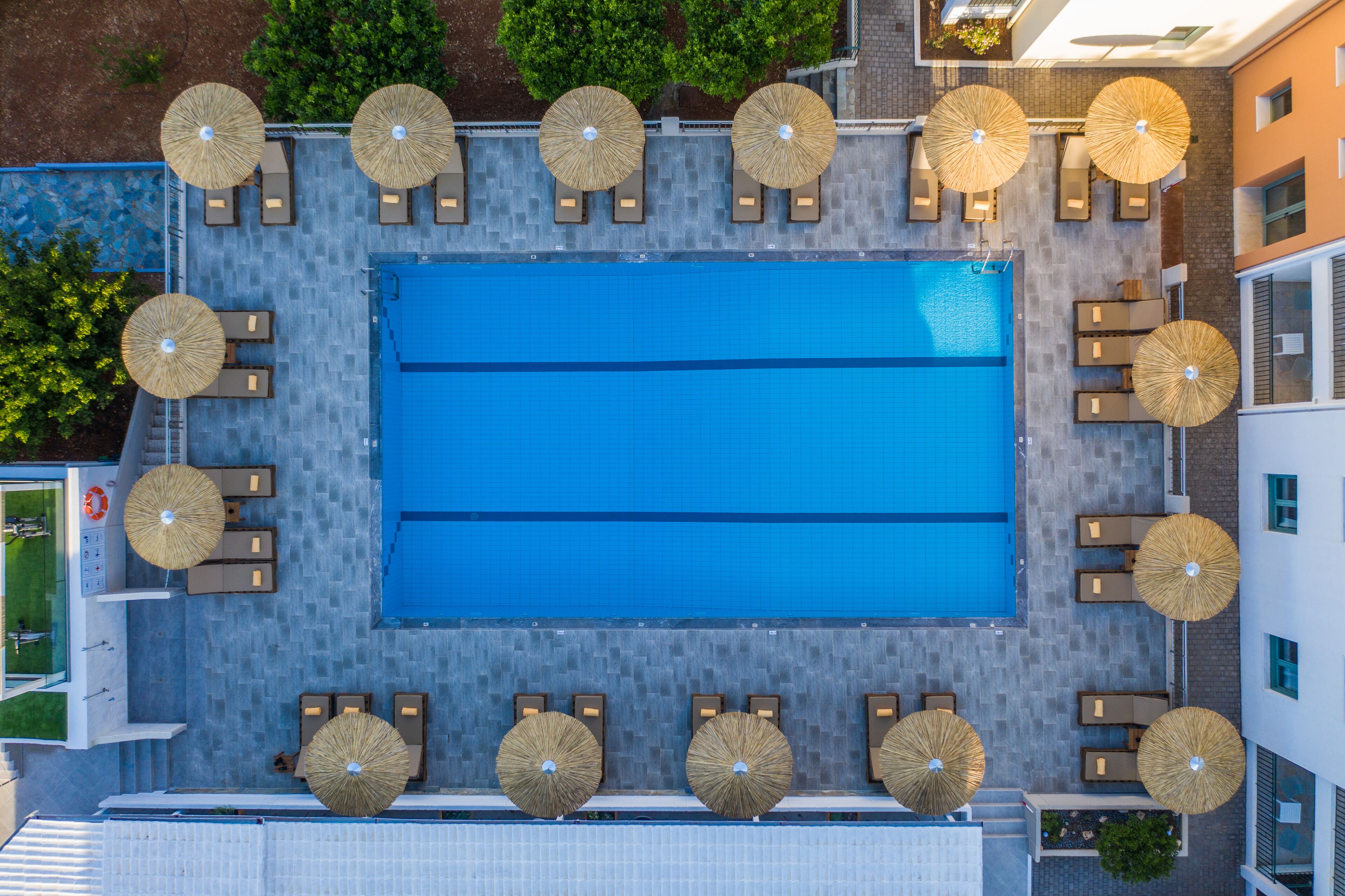 An aerial view of a rooftop pool area with multiple sun loungers and umbrellas, surrounded by a tiled deck and adjacent buildings.
